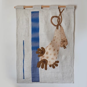 Puede incluir: Colgante de pared textil con un diseño caprichoso. La obra de arte presenta un fondo a rayas beige claro con una franja vertical azul y una forma abstracta marrón moteada. La pieza está montada sobre una barra de madera.
