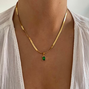Puede incluir: Collar dorado con cadena de espiga y colgante rectangular verde esmeralda. El collar se luce sobre una blusa blanca, creando un contraste que resalta el diseño de la joya.