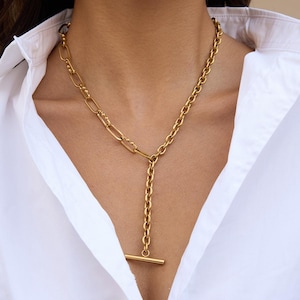 May include: A gold-coloured necklace with a distinctive T-bar pendant. The necklace features a combination of chain styles, including a curb chain and a paperclip chain. The necklace is worn over a white button-down shirt.