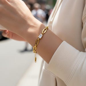 May include: Gold-toned bracelet featuring an oval link chain design with a central square-cut, clear gemstone. The bracelet is shown on a person's arm, with a neutral-toned long-sleeved top in the background. The bracelet's clasp is visible.