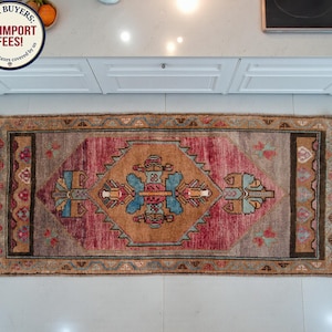 Puede incluir: Alfombra rectangular con un diseño geométrico central en tonos marrón, rojo y azul. La alfombra tiene un borde con un patrón repetido y está colocada sobre un suelo claro. El diseño es simétrico.