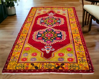 May include: A red, yellow, and green patterned rug with intricate floral designs. The rug has a fringed edge and is laid out on a wooden floor.