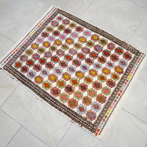May include: A white rug with a geometric pattern in shades of red, green, blue, and yellow. The pattern is made up of small diamonds and squares.