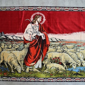 Op de afbeelding: Rood tapijt met een religieuze scène. Een figuur in een rode jurk houdt een staf en een lam vast, omringd door schapen. De rand heeft een decoratief patroon en rode franjes.