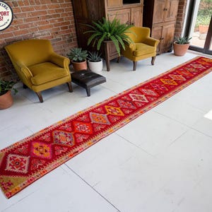 May include: A long, narrow runner rug featuring a vibrant red base with repeating geometric diamond patterns in shades of orange, yellow, pink, and blue. The rug has a distinct border and a textured weave, extending across a white floor surface.