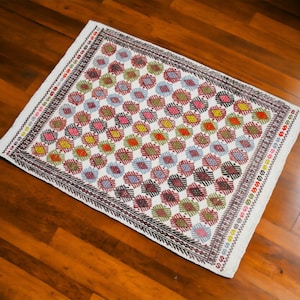May include: A white rug with a geometric pattern of red, blue, green, and yellow diamonds. The rug has a white border with a black and brown trim.