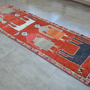 May include: A long, narrow rug with a red background and a colorful geometric pattern. The pattern features stylized figures and animals in shades of orange, blue, white, and black.