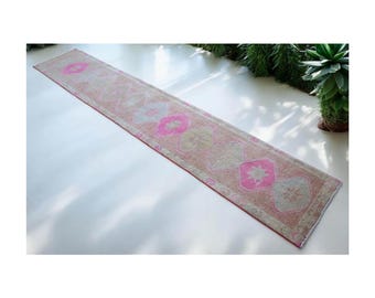 Puede incluir: Una alfombra larga y estrecha de color rosa y beige con un patrón geométrico. La alfombra tiene un borde con flecos y está colocada sobre una superficie blanca.