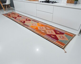 May include: A long, rectangular runner rug with a geometric diamond pattern in shades of orange, red, brown, and gray. The rug is placed in front of a white kitchen cabinet. The rug has a dark brown border and tassels at both ends.