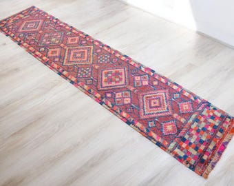 Puede incluir: Una alfombra larga y estrecha con un patrón geométrico en tonos de rosa, rojo, azul y blanco. La alfombra tiene flecos en ambos extremos.