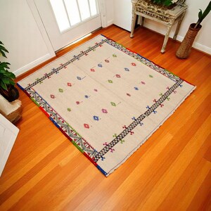 May include: Beige rectangular rug with a geometric border in red, blue, green, and black. The rug has a repeating pattern of small, colorful cross-like shapes. The rug is placed on a hardwood floor.