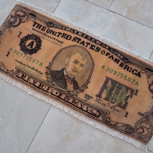 May include: A brown and beige rug with a frayed edge, depicting a five dollar bill. The rug features the text "THE UNITED STATES OF AMERICA" and the serial number "A30973507A".