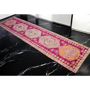 Può includere: Un tappeto da corridoio lungo e stretto con sfondo rosa e intricati motivi geometrici in tonalità beige, arancio e rosa. Il tappeto ha un bordo con frange.