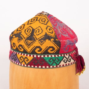 May include: A colorful, patterned hat with a tassel. The hat is made of woven fabric and has a geometric design in shades of red, yellow, green, black, and purple.