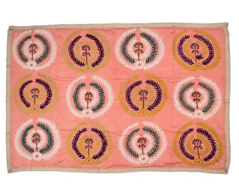 May include: A coral textile with a repeating pattern of embroidered circles. Each circle features a central floral design surrounded by a wreath of leaves in white, green, yellow, and navy blue. The edges are trimmed with a neutral-toned border.
