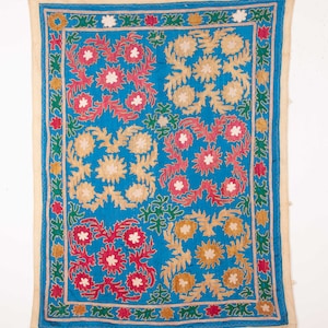 May include: A blue fabric wall hanging with a repeating pattern of embroidered flowers in shades of red, pink, white, and gold. The flowers are surrounded by green leaves and a border of white and gold.
