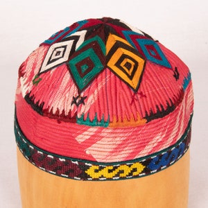 Puede incluir: Un sombrero de tela rojo, rosa y blanco con diseños geométricos bordados de colores. El sombrero tiene una parte superior redondeada y una banda de bordado negro, amarillo y azul alrededor de la parte inferior.