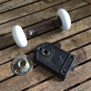 May include: A vintage door hardware set consisting of two white porcelain knobs connected by a dark metal spindle, a circular brass-toned metal rosette with screw holes, and a rectangular black metal mortise lock mechanism with a square hole.
