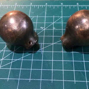 May include: Two spherical, dark brown metal knobs with a smooth, polished finish. Each knob features a tapered base with a small circular opening. The objects are positioned on a green cutting mat with white grid lines and inch markings visible.