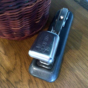 May include: A black and silver stapler with a silver top that says "ARROW 20/6" on it. The stapler is sitting on a wooden surface.