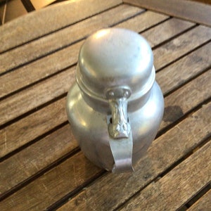 May include: A silver aluminum sugar bowl with a lid and a decorative handle. The bowl is sitting on a wooden table.