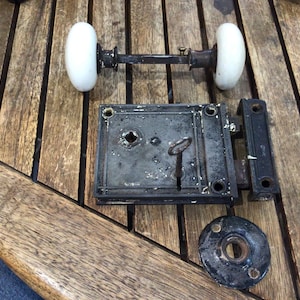 May include: A vintage door hardware set featuring two white porcelain knobs connected by a dark metal spindle, a rectangular black iron mortise lock case with an inserted skeleton key, and a circular black metal escutcheon plate with screw holes.