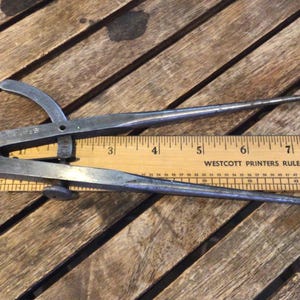 May include: A vintage metal caliper tool with pointed ends, resting on a wooden ruler. The ruler, marked with inches, reads "WESTCOTT PRINTERS RULE". The caliper is open, showing its adjustable design.
