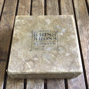 May include: A square, beige-colored box with a textured surface. The center features black printed text that reads: RHODES KRISS KROSS SAFETY BLADE STROPPER, PRICE 50 DOLLARS, MADE BY RHODES MFG CO ST LOUIS USA.