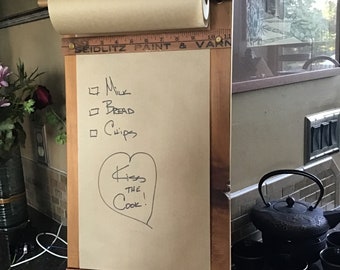 May include: Wooden kitchen notepad with a roll of paper and a pencil holder. The notepad has a list of items to buy and the text "Kiss the Cook!" written on it. The notepad is decorated with rulers and the text "Seidlitz Paint & Varn" and "Lacquers".