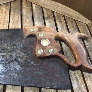 May include: An antique hand saw with a wooden handle and a rusty, toothed blade. The handle is attached to the blade with brass rivets. The saw is resting on a wooden surface.
