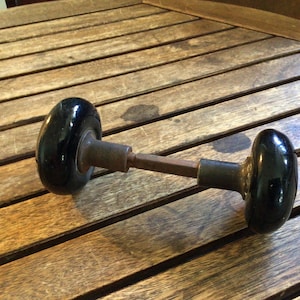 May include: A vintage door handle with two black, oval-shaped knobs connected by a metal rod. The knobs have a glossy finish and are mounted on a wooden surface.