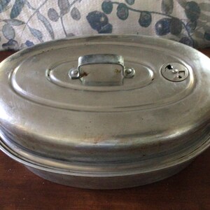 May include: An oval-shaped silver-toned metal roasting pan with a matching lid. The lid features a central arched handle and a small circular adjustable vent. The exterior surface of the metal shows a smooth, reflective finish with minor signs of wear.