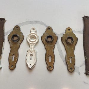 May include: A collection of six metal door plates arranged in a row. Two large rectangular plates with rounded edges flank four smaller, ornate, teardrop-shaped plates. Five plates have a tarnished brass finish, while one central plate is white.