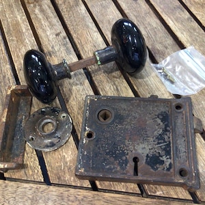 May include: A vintage door hardware set featuring two round black porcelain knobs connected by a metal spindle, a rectangular iron mortise lock box with a keyhole, a circular metal rosette, a metal strike plate, and a small plastic bag with a screw.