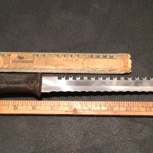 May include: A serrated knife with a dark wooden handle and a metallic blade featuring a notched top edge, positioned above a wooden ruler measuring 16 inches in length, accompanied by a rectangular paper label with vintage-style text.