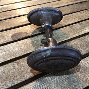 May include: A vintage-style metal door knob set featuring two oval-shaped handles with a dark, weathered patina and a beaded border detail. The two knobs are connected by a central square spindle shaft with a visible adjustment screw on the neck.