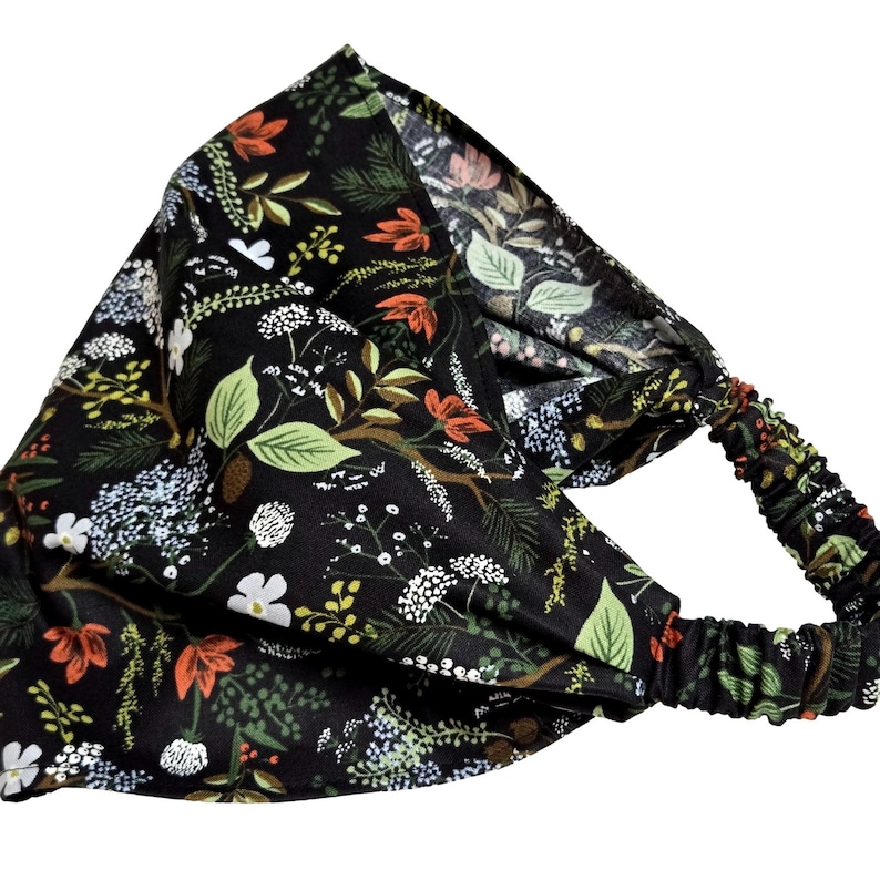 May include: A black headband with an all-over floral pattern. The design features green leaves, white flowers, and orange accents. The elastic band is black with a matching floral print.