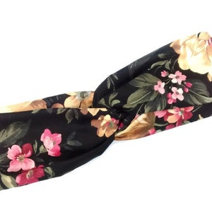 May include: Black floral headband featuring a twisted knot design. The fabric is adorned with a pattern of pink, yellow, and brown flowers with green leaves. The headband is a stylish hair accessory.