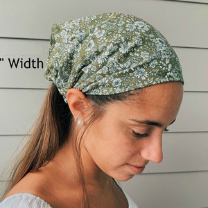 May include: A green floral print head wrap, 10 inches wide, is worn on the head. The fabric features a pattern of small white flowers and green leaves. The head covering is tied at the back of the head.