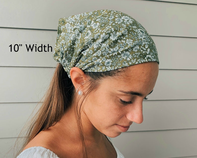 May include: A green floral print head wrap, 10 inches wide, is worn on the head. The fabric features a pattern of small white flowers and green leaves. The head covering is tied at the back of the head.