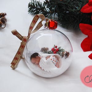 May include: A clear glass ornament with a baby doll inside, wearing a white and red outfit. The ornament is decorated with a red bow and a red bell. The ornament is surrounded by white snow and a small green tree.