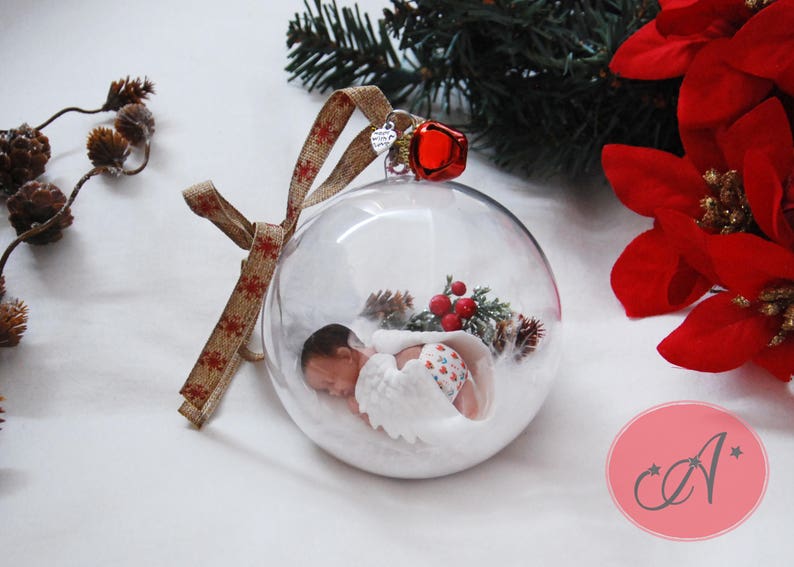 May include: A clear glass ornament with a baby doll inside, wearing a white and red outfit. The ornament is decorated with a red bow and a red bell. The ornament is surrounded by white snow and a small green tree.