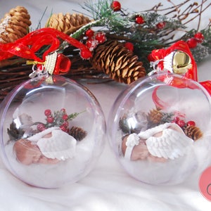 May include: Two clear glass Christmas ornaments with a sleeping baby angel inside. Each ornament is decorated with white faux snow, pine sprigs, and red berries. The ornaments are hanging from red and gold ribbon.