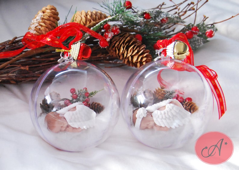 May include: Two clear glass Christmas ornaments with a sleeping baby angel inside. Each ornament is decorated with white faux snow, pine sprigs, and red berries. The ornaments are hanging from red and gold ribbon.