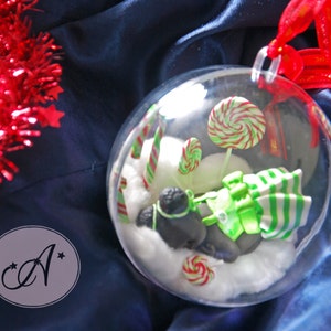 Puede incluir: Una bola de Navidad de cristal transparente llena de caramelos en miniatura, una figura de pingüino y nieve artificial. El adorno presenta piruletas a rayas rojas y verdes, bastones de caramelo y un regalo envuelto. Una cinta roja y oropel añaden acentos festivos.