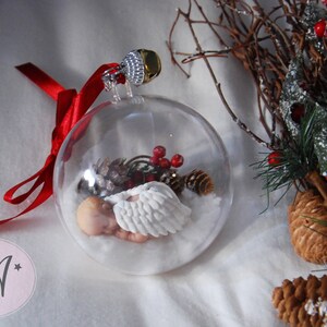 May include: A clear glass Christmas ornament with a sleeping baby angel inside. The angel is wearing a white gown and has a halo. The ornament is filled with white faux snow and red berries.