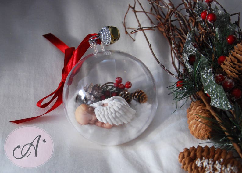 May include: A clear glass Christmas ornament with a sleeping baby angel inside. The angel is wearing a white gown and has a halo. The ornament is filled with white faux snow and red berries.