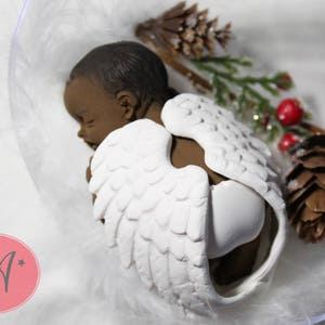 May include: A sleeping baby angel figurine with brown skin and white wings. The figurine is placed in a clear plastic container with white feathers and a small pine cone.