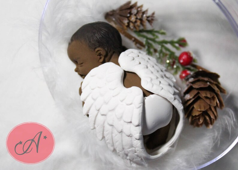 May include: A sleeping baby angel figurine with brown skin and white wings. The figurine is placed in a clear plastic container with white feathers and a small pine cone.