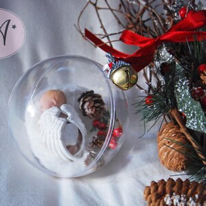 May include: A clear glass ornament with a small, white porcelain angel inside. The ornament is surrounded by pine branches, pine cones, and red berries.